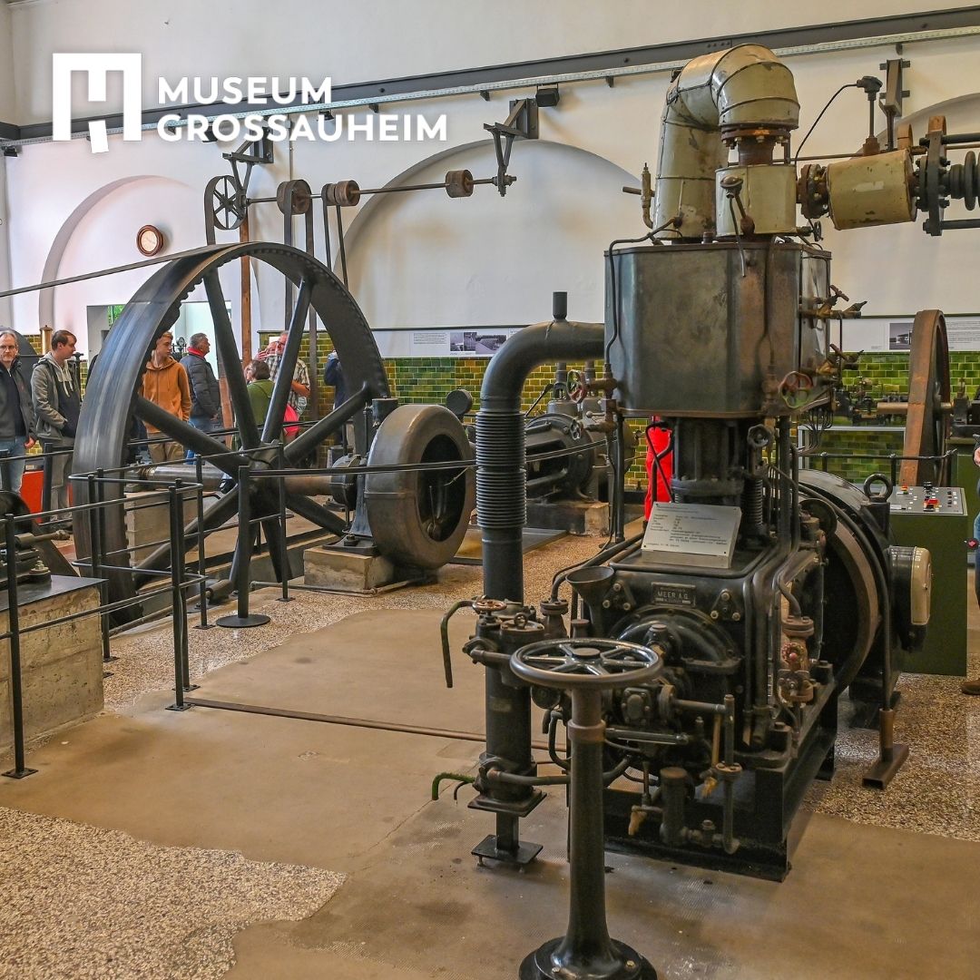 In der Maschinenhalle sieht man verschiedene Dampfmaschinen mit einer Führungsgruppe vor dem Rad der liegenden Dampfmaschine links.
